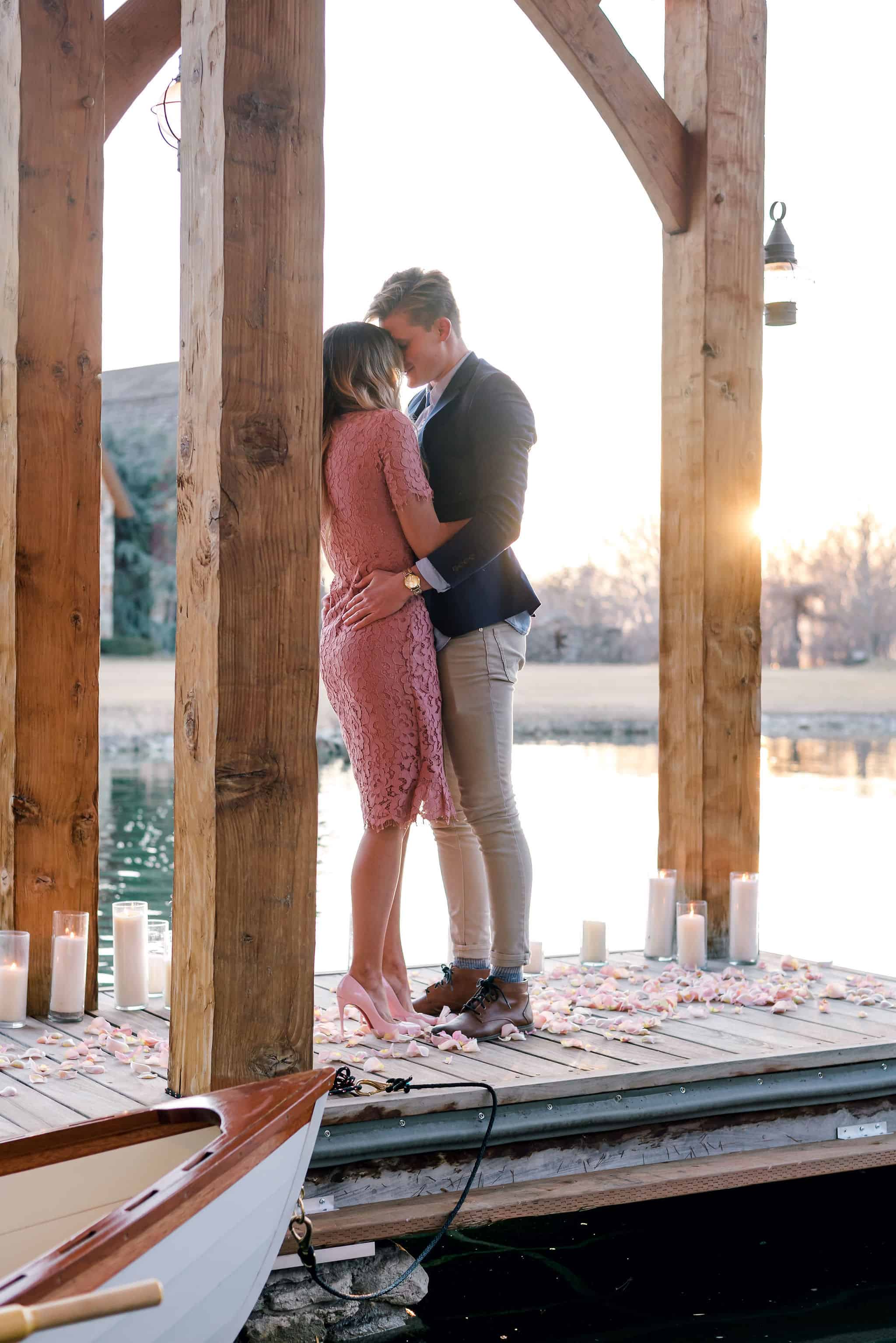Stunning proposal on a dock scattered with flower petals and candles, the couple embrace next to a boat as the sun sets