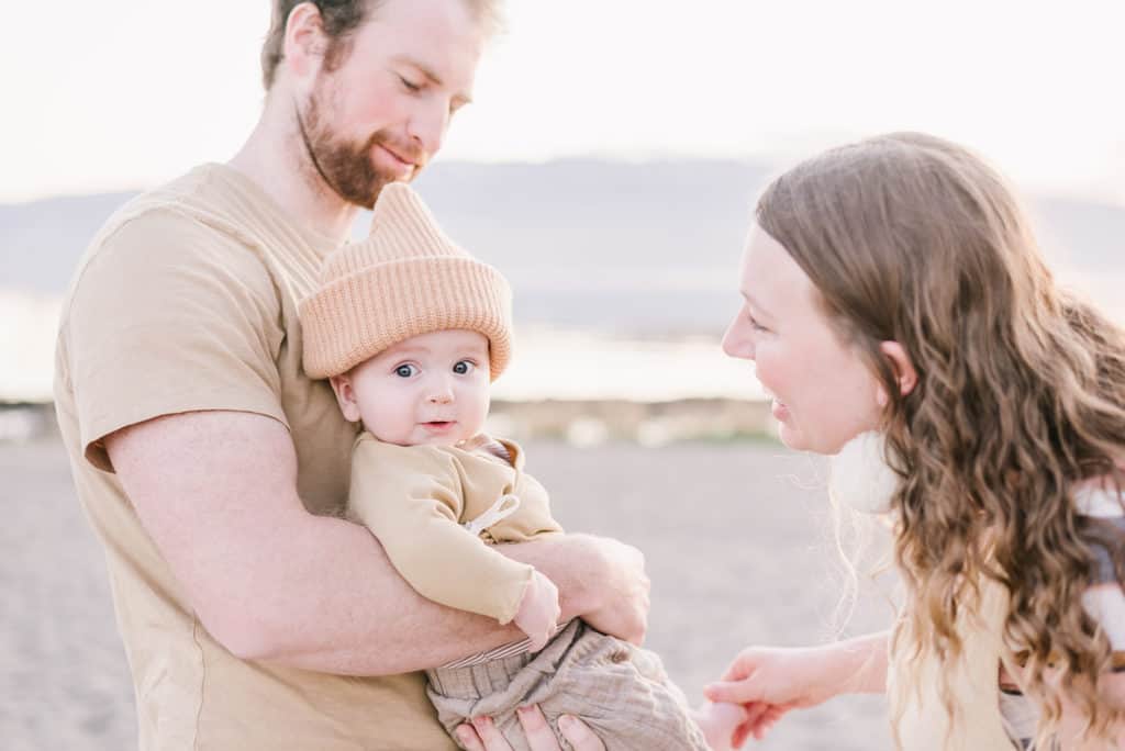 Baby in tan clothes and beanie looks towards the camera as mom and dad play with him in family photography session on Vineyard Beach