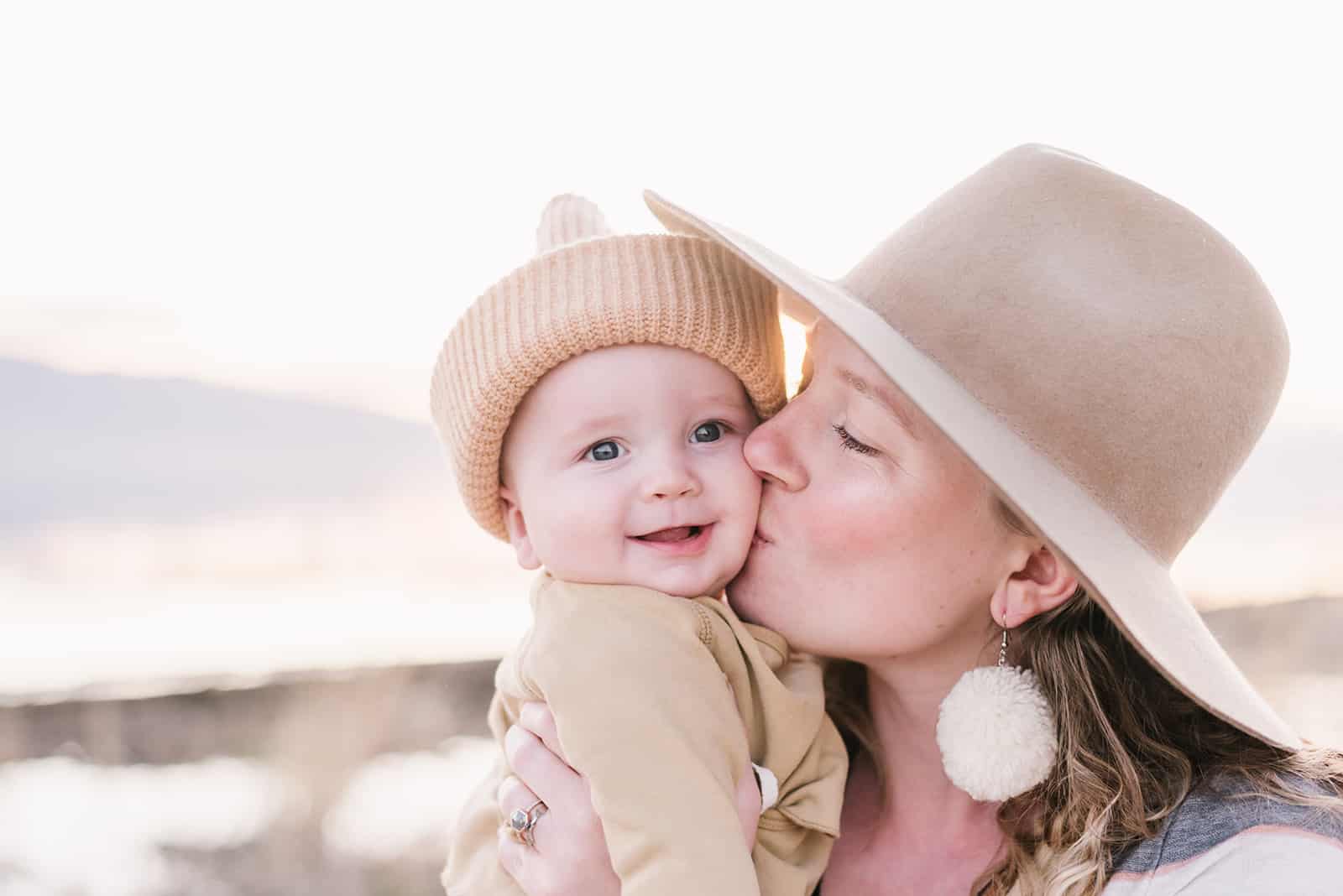 Mom in light brown hat kisses baby in tan front wrap shirt and beanie on vineyard beach during their family pictures.